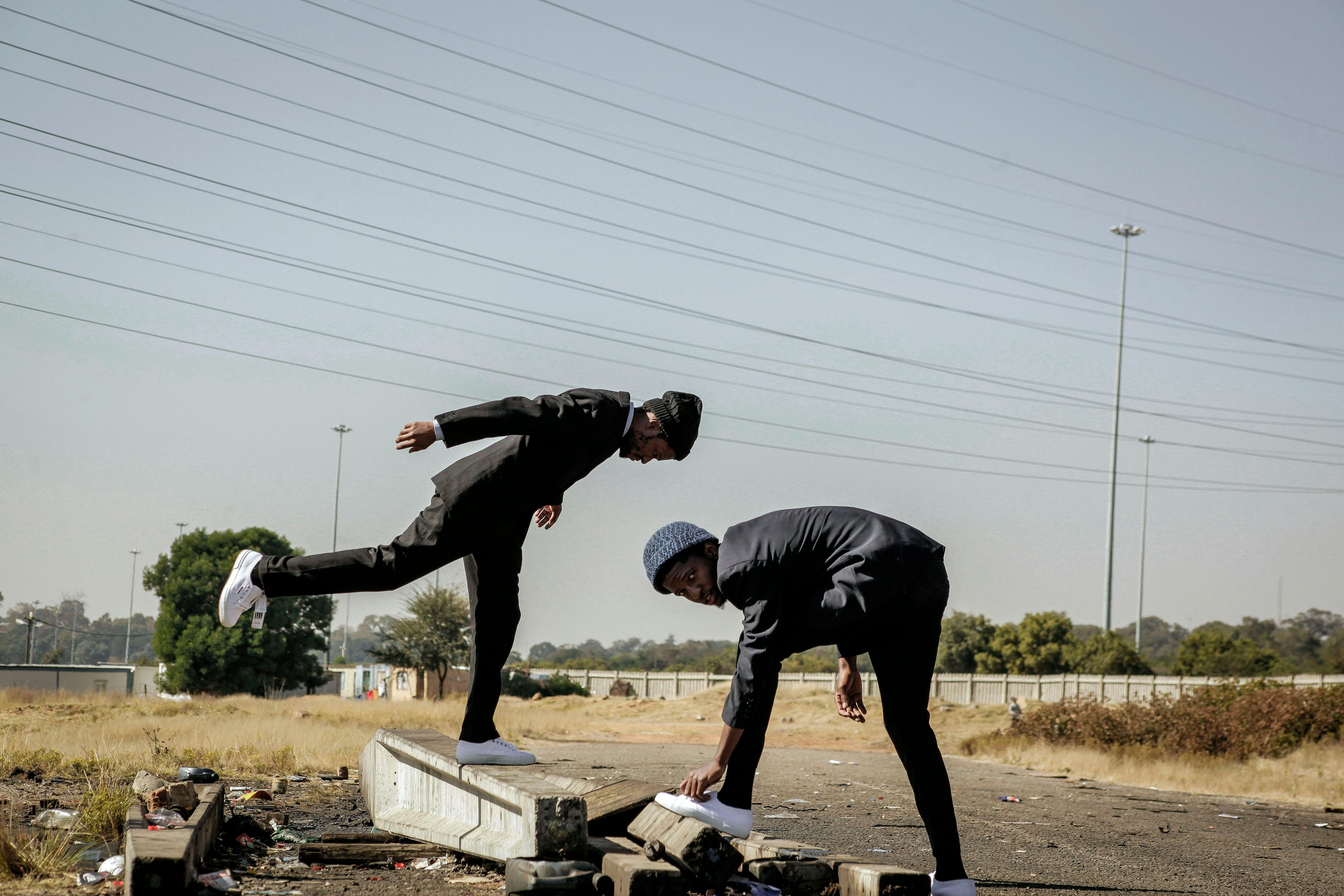 Two men in black suits and beanies balancing and posing on a concrete barrier outdoors.