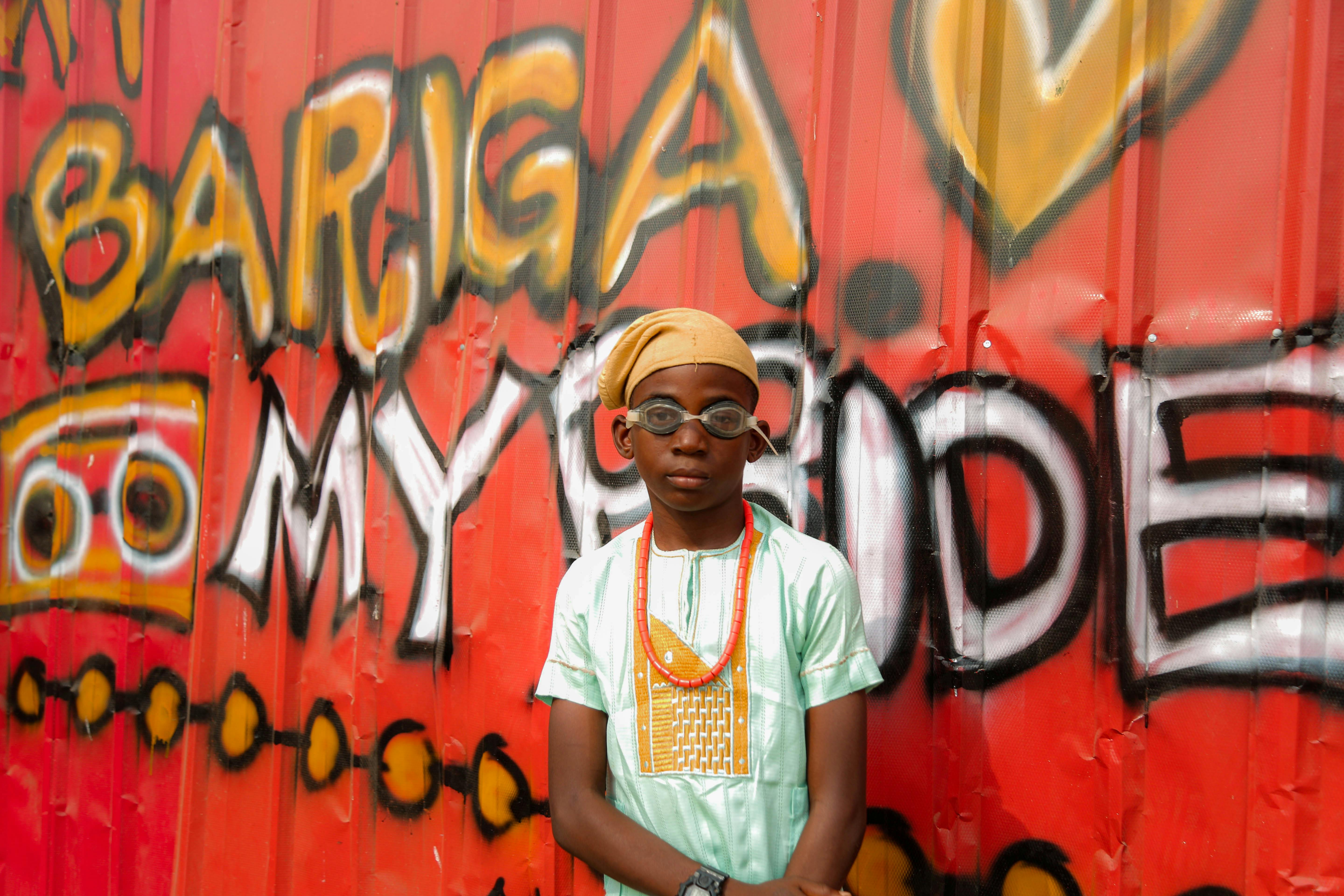 Boy in traditional attire and swimming goggles stands before colorful graffiti wall with 'BARGA MY SIDE'.