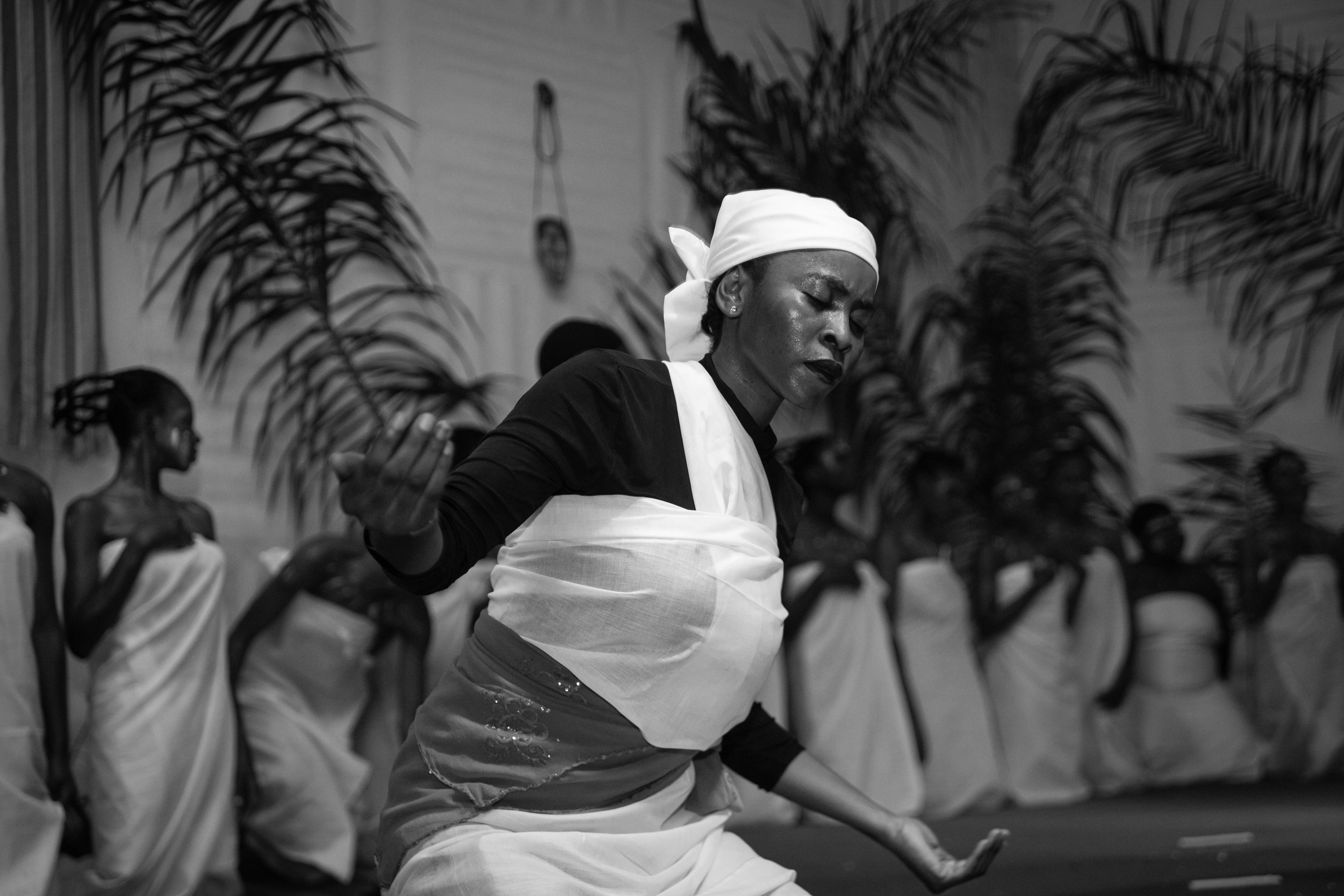 Woman with headscarf dances with eyes closed, others wrapped in cloth stand in background among plants.