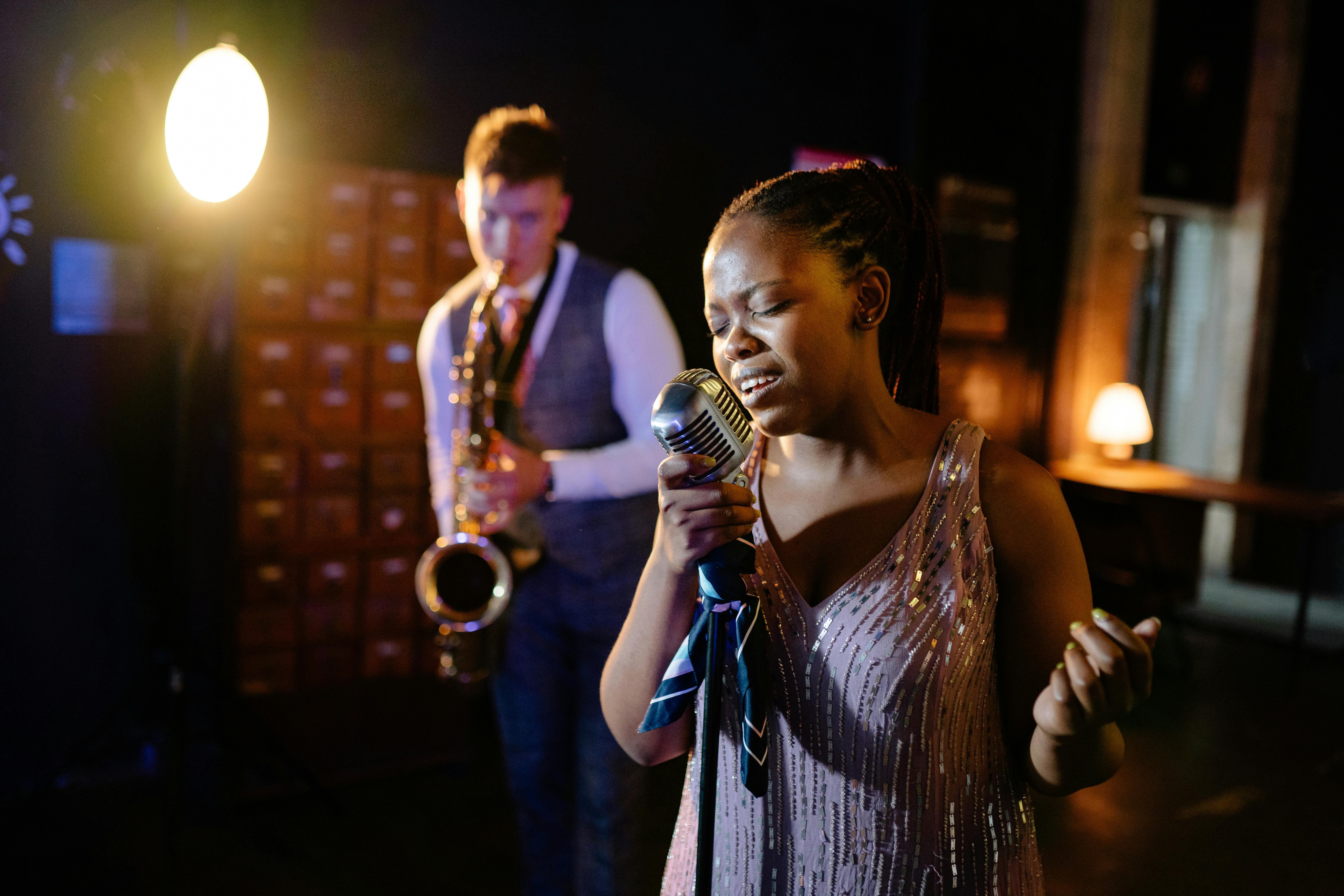 Woman singing into a vintage microphone with a man playing saxophone in a dimly lit room.